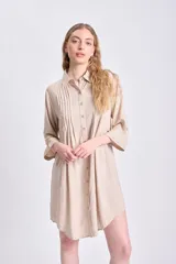 Vestido camisero corto de color beige, con cuello clásico, cierre frontal de botones y detalle de tablas verticales en el frente. Presenta mangas tres cuartos y un corte holgado con ruedo redondeado.