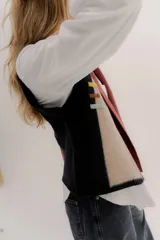 Chaleco abierto de tejido de punto, con diseño de bloques de color verticales en gris claro, negro y gris oscuro. Presenta un cuello ancho tipo chal y un detalle de tres pequeñas aplicaciones bordadas horizontales en tonos verde oliva, naranja quemado y celeste pálido en el frente.