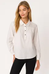Blusa de algodón color blanco, con cuello camisero, cierre frontal parcial con botones y detalle de encaje en la parte superior delantera. Presenta mangas largas con puños elásticos y un corte holgado.