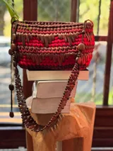 Bolso de mano tejido a mano con tiras de material orgánico en tonos tierra (rojo, marrón, beige) sobre una base de eco-cuero. Posee un asa de mano hecha de un cinturón vintage.