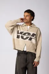 Campera tejida de punto con cierre frontal completo y cuello polo. Presenta un diseño de franja horizontal ancha en color crema con estampado gráfico oscuro sobre una base de color marrón. Posee puños y ruedo acanalados, con un pequeño parche de marca en el puño izquierdo.