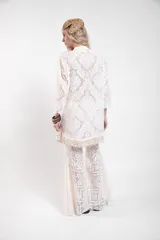 Vestido camisero corto de encaje blanco con diseño ornamental, escote en V con cordón ajustable y ojales metálicos, mangas 3/4 acampanadas y ruedo con detalle de flecos de caracoles tejidos a mano.
