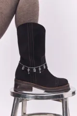 Bota de caña media confeccionada en cuero gamuzado color negro, con costuras a la vista en tono contrastante. Presenta un detalle de cadena metálica plateada con dijes colgantes en forma de llaves y candados alrededor del tobillo. Cuenta con un taco cuadrado de madera y suela de goma.