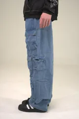 Pantalón tipo cargo de jean en color azul medio, con corte baggy fit y tiro bajo. Presenta múltiples bolsillos laterales con solapa y cierre de botón, además de un diseño holgado con frunces en la parte inferior de las piernas.