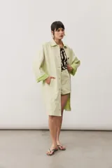 Chaqueta oversize de gabardina color verde claro, con solapa clásica, bolsillos frontales, cierre de botones oscuros y mangas largas con vuelta en tono verde lima vibrante.