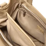 Cartera tipo tote de tamaño pequeño en color beige, confeccionada en material textil. Cuenta con doble asa de mano, correa de hombro ajustable y extraíble, y un detalle decorativo de borlas de cordón en la parte frontal. Incluye un aplique circular con el logo de la marca en el centro.