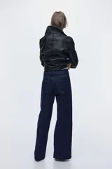 Pantalón de jean de denim azul oscuro, de tiro alto y piernas amplias con corte holgado. Presenta solapas de bolsillo decorativas con botón en el frente y costuras verticales visibles a lo largo de las piernas.