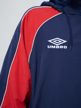 Campera deportiva Umbro del Club Nacional de Football, modelo Venti, para hombre. Diseño en color blanco con mangas azules y detalles de líneas rojas. Cuenta con capucha con cordón ajustable, cierre frontal y bolsillos. Presenta el logo de Umbro y el escudo del club bordados en el pecho.
