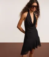 Vestido corto negro de tejido liviano, con escote halter que forma un drapeado (escote degagê) en el busto y ruedo asimétrico con volados.