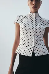 Top de popelín blanco con estampado de lunares negros, de manga corta, cuello redondo y cierre frontal con botones. Cuenta con bolsillos de parche en la parte delantera.