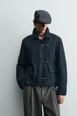 Chaqueta de denim de algodón en color azul oscuro, con cuello solapa y manga larga. Presenta cierre frontal con cremallera oculta bajo una solapa con alamares metálicos, bolsillo de vivo en el pecho y bolsillos de plastrón con solapa en la cadera.