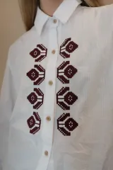 Camisa de algodón con corte clásico, cuello camisero y mangas largas con puños abotonados. Presenta un bordado geométrico en color bordó en la parte frontal y detalle de frunce en la espalda.