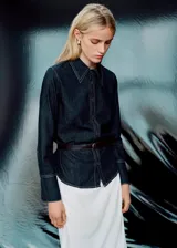 Camisa de denim oscuro con costuras en contraste, cuello camisero clásico, cierre frontal con botones y mangas largas con puños abotonados.