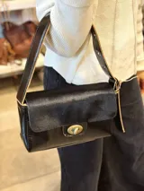 Cartera de cuero color negro, de diseño rectangular con solapa de pelo vacuno en la parte superior, cierre frontal de giro metálico en tono bronce y correa regulable para el hombro.
