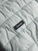 Campera acolchada de poliéster reciclado, color gris claro, con cuello alto y cierre frontal de cremallera negra. Presenta costuras horizontales y una insignia rectangular con el logo de Calvin Klein en el pecho.