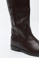 Bota de caña alta estilo western confeccionada en cuero sintético, con bordados decorativos en relieve a lo largo de la caña y puntera afinada.