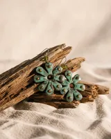 Aros colgantes con diseño de flor de ocho pétalos en cobre oxidado con pátina turquesa y detalles de tachas metálicas. Incluye pasante de plata esterlina y una pequeña cuenta de cristal verde en la parte superior.