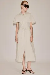 Vestido camisero midi de algodón con diseño de capa, cuello solapa, manga corta tipo capa y trabillas en los hombros. Presenta cierre frontal con botones, bolsillos delanteros y cinturón ajustable del mismo tejido con lazada. El bajo tiene un acabado en línea evasé.
