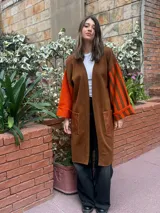 Saco largo de lana merino en color marrón, con diseño abierto sin botones. Presenta mangas anchas con estampado de rayas verticales en tonos naranja y marrón, y un bolsillo frontal aplicado en material símil cuero con textura.