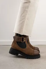 Bota tipo Chelsea de cuero marrón con suela track negra de goma. Presenta paneles elásticos laterales y una costura reforzada en la puntera. Incluye una correa de cuero desmontable con hebilla metálica.