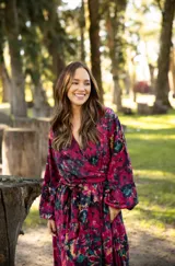 Vestido largo de lino con escote cruzado, mangas largas abullonadas y lazo ajustable en la cintura. Presenta un estampado floral vibrante en tonos fucsia, violeta y verde.