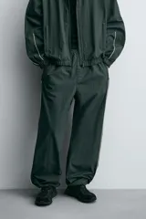 Pantalón jogger de corte relajado confeccionado en tejido técnico de color verde oscuro. Presenta cintura elástica con cordón interior, bolsillos laterales con cierre y bajo ajustable con elástico. Incluye detalles de líneas en contraste a lo largo de las piernas.