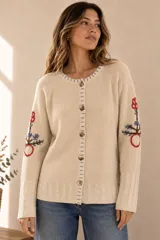 Saco de punto color beige, confeccionado en algodón, con cuello redondo, mangas largas y cierre frontal con botones. Presenta detalles bordados con diseño floral en las mangas y ribetes con rayas azules y blancas en la botonera y el cuello.