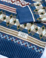Buzo tejido de punto con diseño de patrones geométricos estilo nórdico en tonos azules, marrones, blancos y verdes. Presenta cuello redondo, puños y ruedo acanalados, con etiqueta de la marca Denali en el pecho.