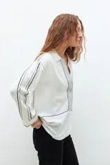 Blusa color crudo de corte recto, con cuello camisero, mangas largas y bordados en contraste de color negro.