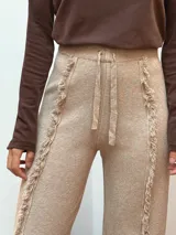 Pantalón de punto color beige, de corte recto y holgado, con cintura elástica y cordón ajustable. Presenta detalles de costuras con flecos verticales a lo largo de las piernas.