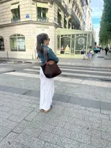 Bolso tipo shopper de cuerpo plegable en color crema o blanco roto, con asas y solapa de cierre en cuero marrón texturizado. Presenta un bolsillo exterior frontal y cierre principal con broche metálico.