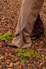Pantalón de sastrería con diseño bicolor asimétrico, combinando una pierna en color marrón oscuro y la otra en beige, ambos con un sutil estampado de rayas diplomáticas. Presenta una silueta de corte recto y relajado, con cierre frontal y botón de nácar.