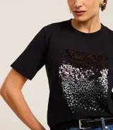 Remera negra de algodón con corte slim, cuello redondo y mangas cortas. Presenta un bloque rectangular frontal con lentejuelas plateadas.