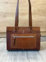 Cartera de cuero marrón de tamaño medio con diseño estructurado, que combina cuero liso y un panel frontal texturizado tipo cocodrilo con bolsillo exterior con cierre. Cuenta con doble asa de hombro y costuras visibles de contraste.