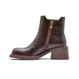 Bota tipo chelsea de cuero color marrón oscuro con acabado desgastado. Presenta paneles elásticos laterales decorados con tachas metálicas doradas, puntera cuadrada y taco bloque de altura media con textura amaderada.