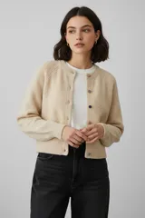 Cardigan de punto color beige, de corte holgado y diseño corto. Presenta cuello redondo, cierre frontal con botones de distintos colores y mangas largas con puños acanalados.