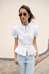 Camisa blanca de manga corta abullonada, con cuello mao y cierre frontal con alamares. Presenta corte evasé con detalle de volados en el bajo.