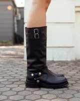 Bota de caña alta confeccionada en cuero negro, con suela track de goma. Presenta un diseño versátil con dos hebillas ajustables en la parte superior de la caña y una correa decorativa con tachas metálicas y argollas en la zona del tobillo.