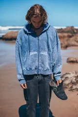 Campera con capucha y cierre frontal doble, confeccionada en tela con efecto lavado azul. Presenta bordado frontal con la frase 'GLASSY WAVES' distribuida sobre los bolsillos tipo canguro.