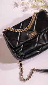 Bandolera negra acolchada con diseño de rombos, cierre dorado y correa de cadena dorada entrelazada con cuero sintético negro.