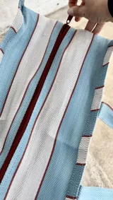 Bolso tipo tote grande con estampado a rayas verticales en tonos celeste, blanco y bordó. Tiene doble asa corta de mano y cierre superior.