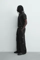 Pantalón cargo de corte relajado en color negro con efecto lavado. Presenta bolsillos laterales tipo cargo con solapas y cintas ajustables, bolsillos frontales y traseros, y detalles de argollas metálicas. Cierre frontal con cremallera y botón.