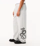 Pantalón de corte super baggy confeccionado en tejido de algodón y poliéster, color gris melange. Presenta un diseño urbano con estampado de serpiente tribal en color negro en las piernas, cintura elástica y bolsillos laterales.