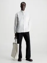 Camisa blanca de manga larga con cuello clásico, cierre frontal con botones, bolsillo de parche en el pecho y logo CK bordado en el bolsillo.