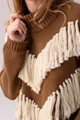 Sweater de punto color crema con cuello medio alto y silueta relajada. Presenta un diseño frontal con líneas en V decoradas con flecos largos de color marrón aplicados a mano.