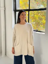 Sweater de punto color beige, de corte holgado y diseño oversize. Presenta cuello redondo, mangas largas con puños ajustados y dos bolsillos frontales tipo parche. El diseño incluye una costura vertical central en el frente y terminaciones acanaladas en el ruedo.