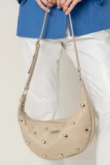 Cartera bandolera de cuero en color beige, con diseño de media luna y tachas metálicas circulares distribuidas en el frente. Cuenta con correa regulable, cierre superior y bolsillo trasero con cierre.