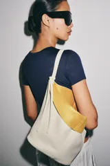 Bolso de hombro de piel en color blanco, con diseño tipo bucket, asa fija y detalle de tira colgante con nudo en el lateral.