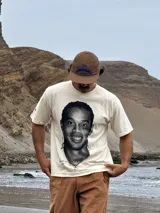 Remera de algodón color beige con estampa fotográfica frontal de Ronaldinho en blanco y negro. Presenta un corte boxy oversize, cuello redondo y mangas cortas.