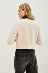 Saco corto de tejido tipo boucle en color crudo, con cierre frontal mediante tres trabas de cuero sintético negro y botones esféricos. Presenta ribetes en contraste negro a lo largo de la abertura frontal y el cuello redondo.